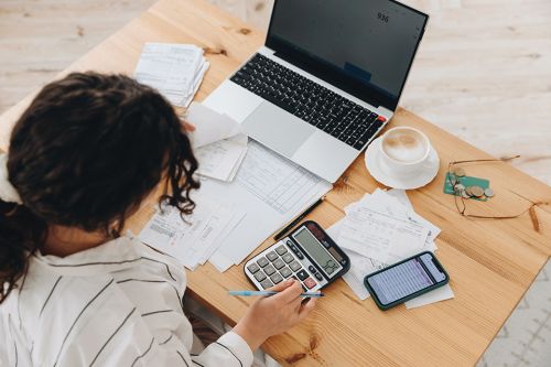 © Sheremetio - stock.adobe.com Foto: Frau tippt auf Taschenrechner mit Laptop am Arbeitstisch